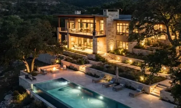 An architectural modern villa in the Texas Hill Country surrounded by mature live oaks and limestone terraces with warm ambient outdoor lighting…